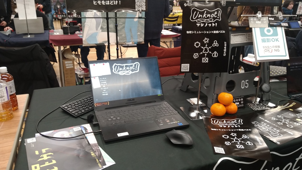 A photograph of an exhibition table. In the table there is a laptop showing the game "unknot!", and several posters, pamphlets, and business cards of the same game