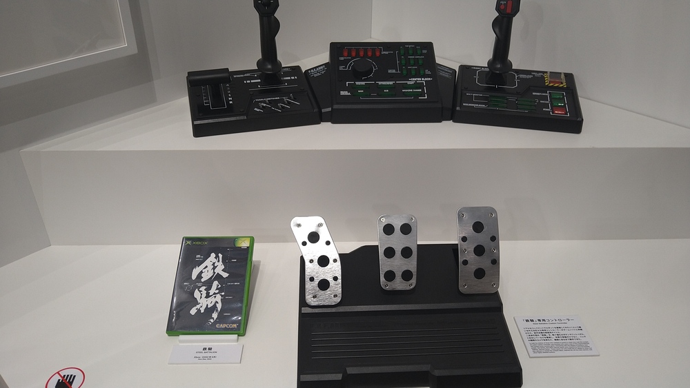 A picture of a game box next to a large controller with two joysticks, three foot pedals, and many buttons, simulating a mecha cockpit.